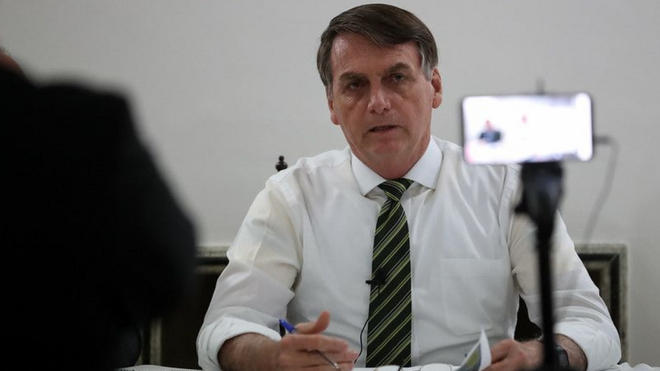 Bolsonaro, sentado diante de mesa, fala para câmera de celular