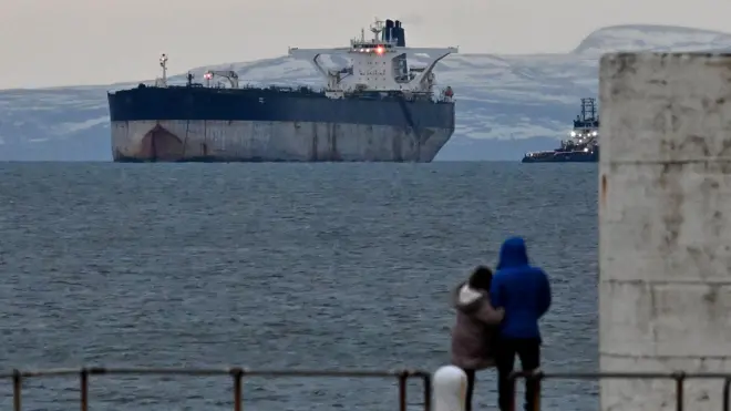 Пара в теплых куртках рассматривает с берега танкер Marinera в холодном шотландском заливе на фоне заснеженных холмов