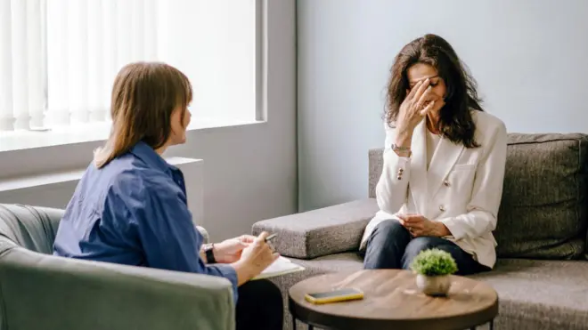 Mujer sentada con la mano en el rostro, afligida por la pena, conversa con una terapeuta que toma nota de la conversación