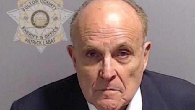 A police mugshot showing Rudy Giuliani in a blue suit and red and blue striped tie which appears to be based on the Stars and Stripes