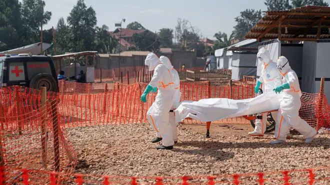 Abakora mu buvuzi batwaye umurambo w'umuntu wishwe na Ebola bawujyanye mu buruhukiro bw'ikigo nderabuzima cya Nyankunde, i Bunia, mu ntara ya Ituri, ku wa gatandatu ushize, kugira ngo benewabo bawusezereho bwa nyuma. Bambaye imyenda yabugenewe y'umweru ibapfutsemo umubiri wose, ndetse n'ingofero z'umweru, banambaye udupfukantoki hamwe n'inkweto. Barimo kugenda mu mbuga y'ivuriro irimo amabuye, ikikijwe n'inzitiro z'ibara rya 'orange'. Ifoto yo ku itariki ya 13 Kamena (6) mu mwaka wa 2026.