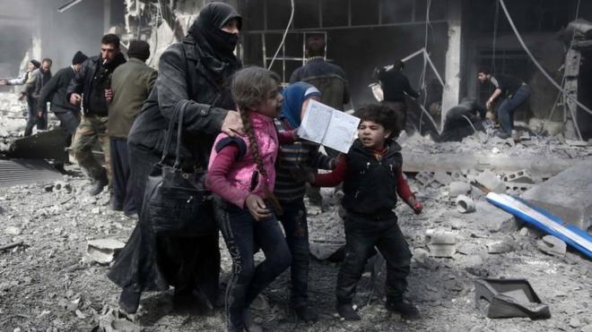 A Syrian woman and children run for cover amid the rubble of buildings following government bombing in the rebel-held town of Hamouria