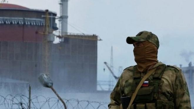 A masked man with an assault rifle stands in front of the nuclear power plant