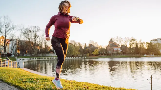 A woman takes a run next to a pond in a UK town. She is looking at her watch and there is early morning light on her face