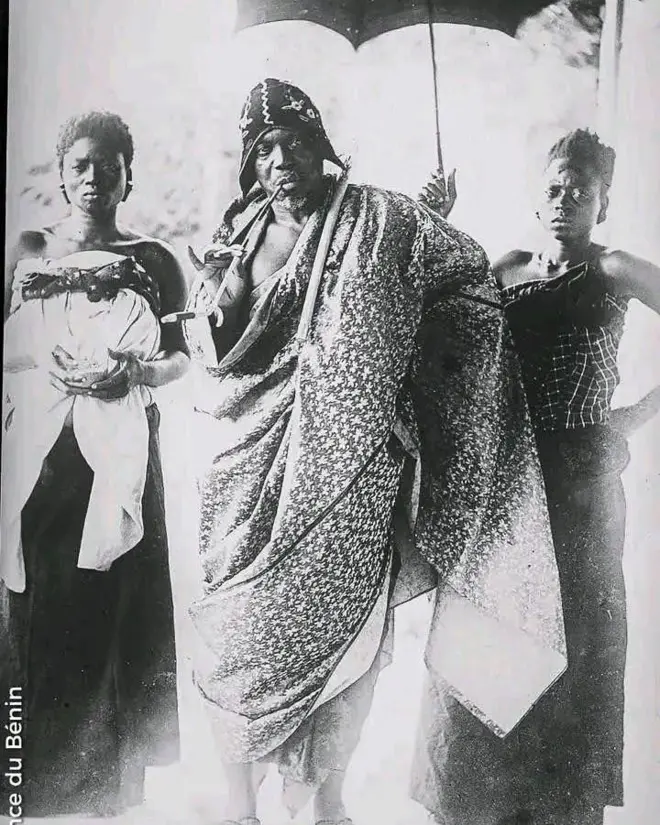 Photo archives du roi Behanzin portant un grand pagne autour de la taille avec une longue pipe entre les lèvres. Derrière lui se trouvent deux femmes dont l'une tient une parapluie pour protéger le roi.