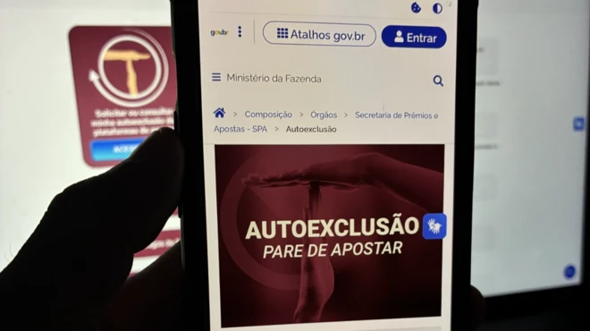 Foto mostra uma mão segurando um telefone celular contendo uma imagem do sistema de autoexclusão para apostadores do governo federal. A imagem é no contraluz e mostra a tela do celular com um ícone vermelho 