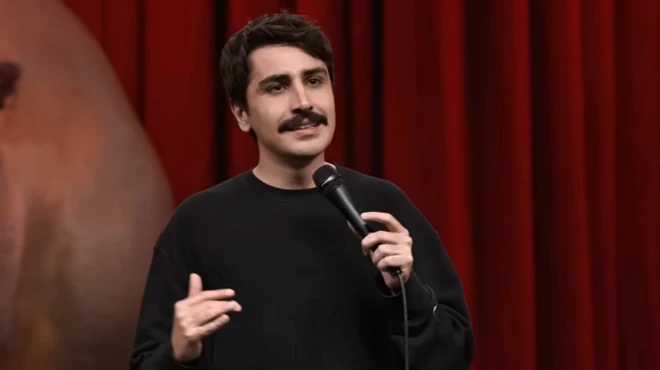 Deniz Göktaş'ın Harbiye'de sahnelediği gösteri sırasında kırmızı bir perde önünde konuşuyor. Bıyıklı ama genç bir görüntüsü var. Bir elinde mikrofon tutuyor.
