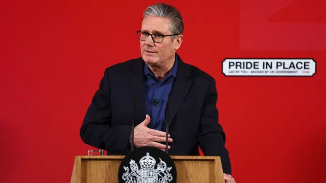 Keir Starmer kırmızı bir arka plan önünde bir kürsüde konuşma yapıyor, duvarda bir sokak ismi tabelası şeklinde üzerinde "pride in place" (yaşanılan yerle duyulan gurur) yazılı bir duvar resmi var. 