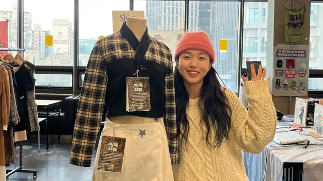 Aktywistka Lee So-yeon macha i ma na sobie różową czapkę oraz kremowy sweter z warkoczowym splotem, stojąc obok manekina ubranego w białą spódnicę i spódnicę w kratkę.