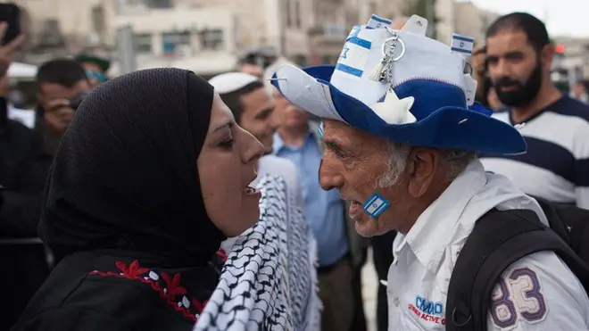 Uma mulher muçulmana e um homem com a bandeira de Israel na bochecha discutem frente a frente 