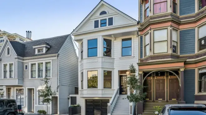 Três casas em uma rua residencial em San Francisco. As casas de três andares parecem limpas e bem conservadas, com plantas e árvores bem aparadas na parte externa.