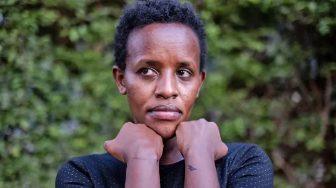 Agather Atuhaire ahagaze yambaye umupira w'amaboko magufi w'umukara urimo utudomo tujya kuba umweru, agaragaza inkovu ku maboko yombi iruhande rw'aho ibiganza bitangirira, nyuma yo kurekurwa n'abategetsi ba Tanzania. Ifoto yo ku itariki ya 23 Gicurasi (5) mu mwaka wa 2025, ari hanze y'ibitaro by'i Kampala, muri Uganda. 