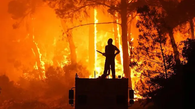 3 Temmuz'da Bucak'daki yangınlara müdahale eden bir ekip üyesi. Arkada alevler görünüyor. Ekip üyesi bir aracın üstünde duruyor ve elinde hortum tutuyor.