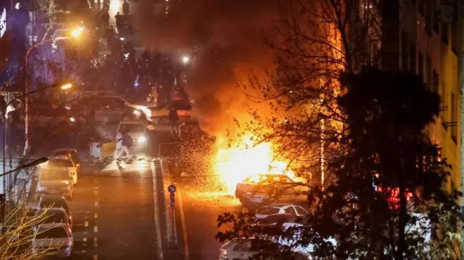 夜の道路に大勢が立つなか、燃える車両からオレンジ色の炎と黒煙が上がっている
