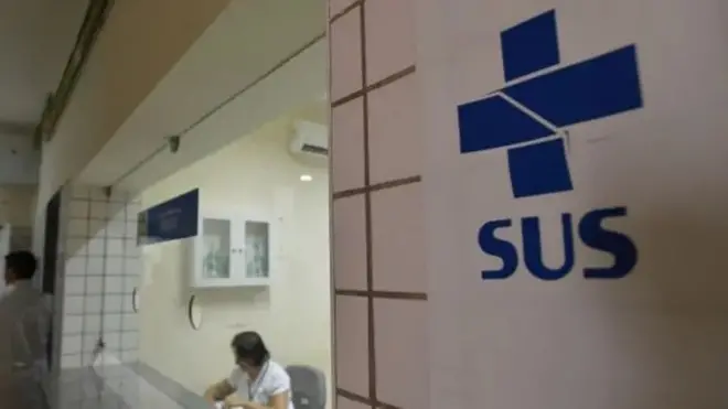 Placa do SUS e ao fundo uma mulher trabalhando