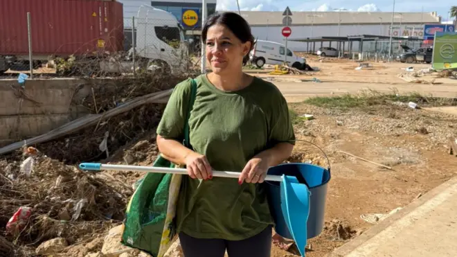 Fotografia colorida mostra mulher brancasambafoot apostascabelo preto carregando pá e baldesambafoot apostasuma rua 
