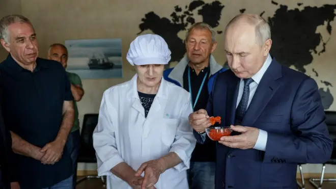 Президент РФ Владимир Путин в темно-синем костюме и синем галстуке держит в руке тарелочку с икрой и ложку, полную красной икры. Рядом женщина в белом колпаке и белом халате. На заднем плане немолодой мужчина в темном поло и джинсах сосредоточенно наблюдает за процессом