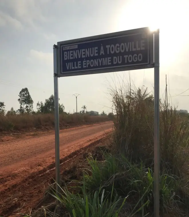 Une plaque indicatif sur laquelle est écrit : "Bienvenue à Togoville, ville éponyme du Togo". Elle est mise sur une piste en latérite avec la lumière du soleil au-dessus de la plaque.