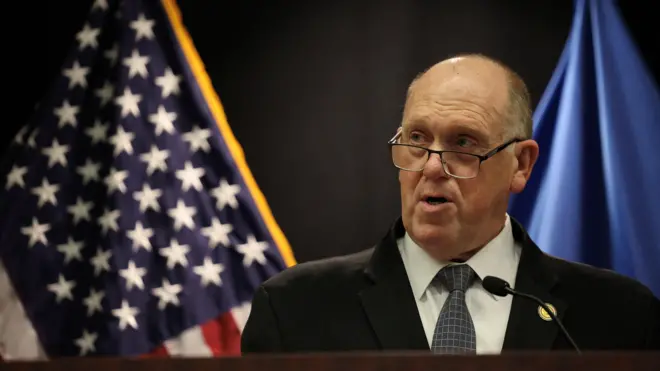 Том Хоман, пожилой лысый мужчина, на фоне американского флага.