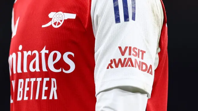 Umwenda wa Arsenal w'umutuku n'umweru wanditseho amagambo Visit Rwanda ku kaboko