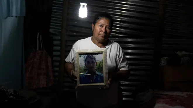 Una mujer sostiene el retrato de su hijo adulto. Está en un cuarto de una casa humilde, a media luz.