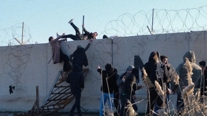Yaklaşık 10 kişi bir duvarın kenarında duruyor. Aralarından üçü tırmanmayı başarmış bir kişi karga tulumba karşı tarafa kendini atarken görünüyor.