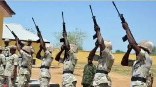 Ọpọ awọn osisẹ ologun na duro wa wa wa ninu asọ ologun alawọ ewe, wọn na ibọn wọn soju ọrun.