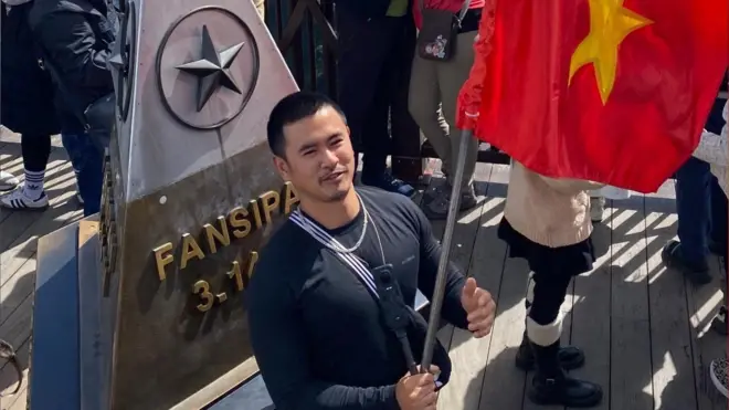 Ike is holding the Vietnamese flag on a balcony. Other tourists are surround Ike - they are taking photos of the view. Ike is wearing a black top with a necklace. Ike had a pyramid shaped statue behind him. The statue has a star on it. 