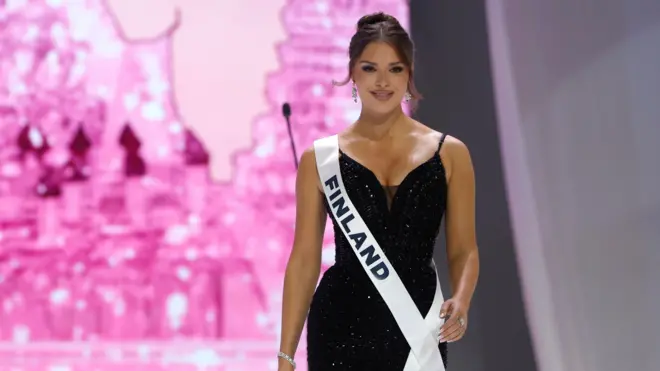 Ciemnowłosa kobieta w czarnej błyszczącej sukience z białym pasem z napisem Finlandia przewieszonym przez ramię i klatkę piersiową