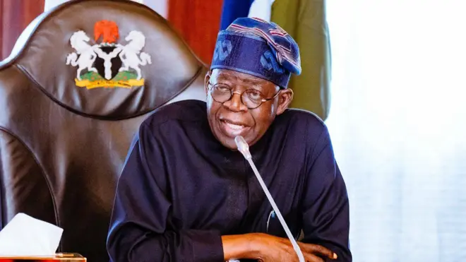 Ààrẹ Tinubu wọ aṣọ dúdú pẹ̀lú fìlà. Gbohùngbohùn wà ní iwájú rẹ̀.