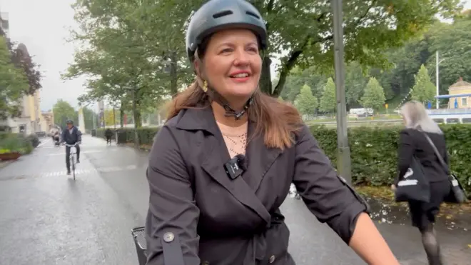 الینا رانتانن در حال دوچرخه‌سواری در کرانه رودخانه، یک بارانی تیره به تن دارد و کلاه کاسکت سیاه رنگ و لبخند می‌زند