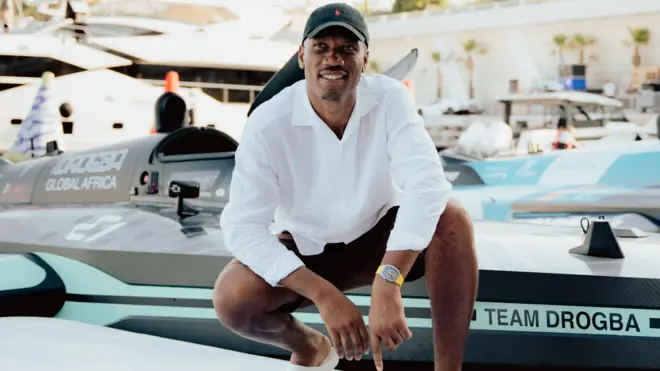 Didier Drogba, vêtu d'un pantalon noir, d'une chemise blanche ample et d'un t-shirt noir, s'accroupit pour poser devant un bateau à moteur noir orné de détails bleu clair et arborant le slogan « Team Drogba » sur sa proue. Dans un décor de marina, plusieurs autres bateaux, dont un très grand yacht, sont visibles à l'arrière-plan.