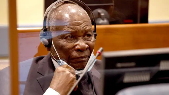 Félicien Kabuga yicaye mu ntebe mu rukiko amanuye agapfukamunwa k'ubururu bwerurutse n'umweru, ku matwi yambaye ibikoresho byo kumufasha kumva ibivugwa (écouteurs), yambaye ikositimu y'umukara ijya kuba ikigina. Afite umusatsi w'imvi n'uruhara. Ifoto yo ku itariki ya 8 Kanama (8) mu mwaka wa 2023, i La Haye (The Hague) mu Buholandi. 