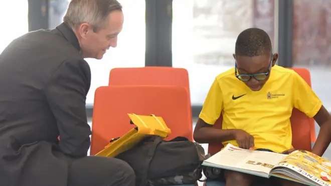 John Todd, head of Charterhouse Nigeria, wey wear grey suit lean forward for one chair near one schoolboy wey wear glasses, and yellow Charterhouse Lagos sports shirt, wey dey read one science book