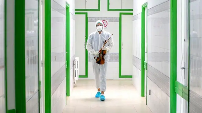 Homem com macacão caminha com violino em corredor de hospital