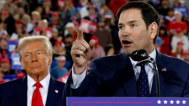 Marco Rubio discursando com Trump atrás, com olhar sério