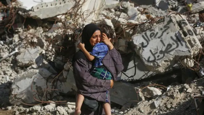19 सितंबर को ग़ज़ा के बुरेजी रिफ़्यूजी कैंप पर इसराइली हमले के बाद एक ध्वस्त हो चुकी इमारत के सामने अपने बच्चे के साथ खड़ी महिला