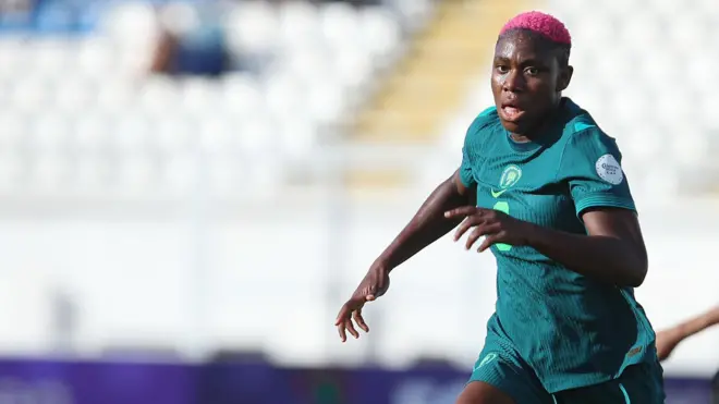 Asisat Oshoala, wearing an all-green Nigeria kit with number 8 on am, dey chase a ball during a match against Zambia, with three opponents in copper jerseys and socks and black shorts seen for di background on di green pitch. An empty stand with white seats dey blurred for di background