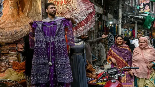 Shopkeeper showing a dress while two women are passing by.