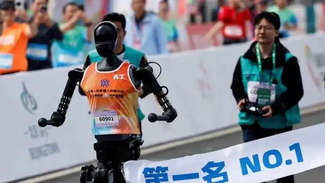 Робот движется по трассе, его фотографируют, сзади стоит человек в салатовом жилете с пультом.