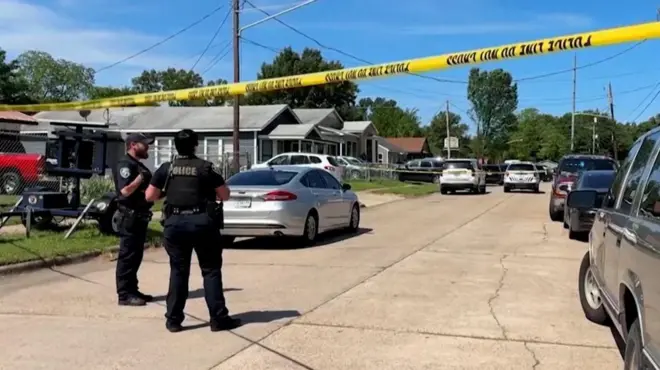 Two police officers stand for street wey don close wit yellow police tape. Cars line di street in front of homes in Shreveport, Louisiana, U.S. on 19 April, 2026, for one still image from video.