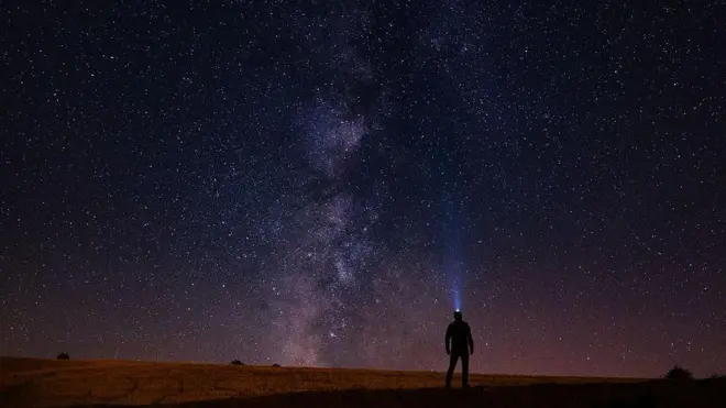 一个人的身影,头戴手电筒,仰望着浩瀚的夜空,布满了无数的星星,中间是银河。