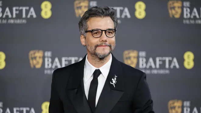 O ator Wagner Moura posando no tapete vermelho do Bafta. Ele veste um terno preto clássico com lapela acetinada, camisa branca e gravata preta. Usa óculos de armação escura e tem barba e cabelos grisalhos bem aparados. Na lapela, há um broche prateado em formato floral. Ao fundo, aparece o painel oficial do evento com o logotipo do BAFTA e a máscara dourada característica da premiação.