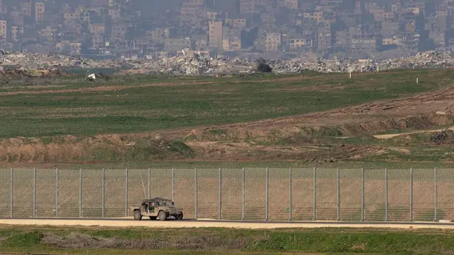 Gazze'nin uzaktan görünümü. Tellerin arkasında enkazlar, önünde bir tank var.