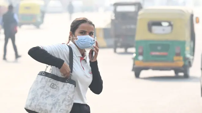 一名戴著口罩、在諾伊達（Noida）街頭邊走邊打電話的女性，背景可見行駛中的三輪嘟嘟車。攝於2025年11月。