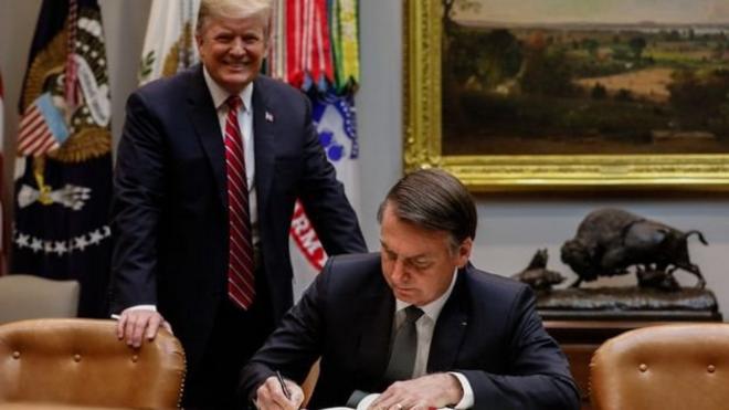 Trump sorrindo, de pe atras da cadeira onde Bolsonaro esta sentado assinando um documento