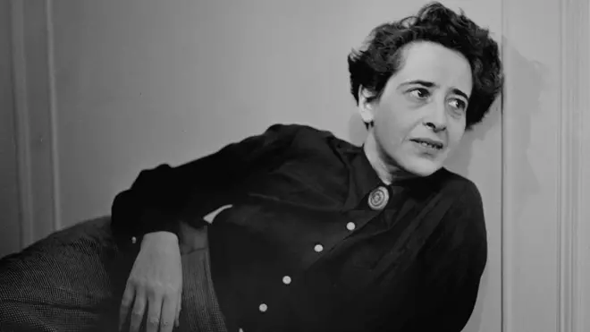 Hannah Arendt con una blusa oscura está recostada sobre una pared en un sofá