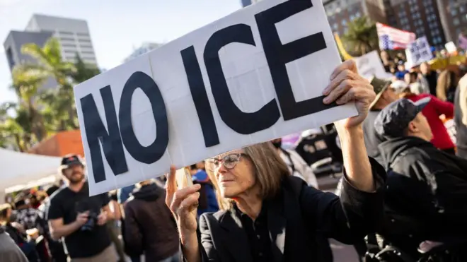 A woman in a dark jacket and wearing glasses holds a placard reading 'NO ICE'. Behind her are other people.