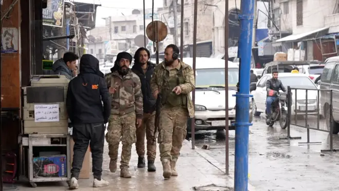 ثلاث مسلحين بلباس عسكري يمشون على الرصيف في أجواء ماطرة