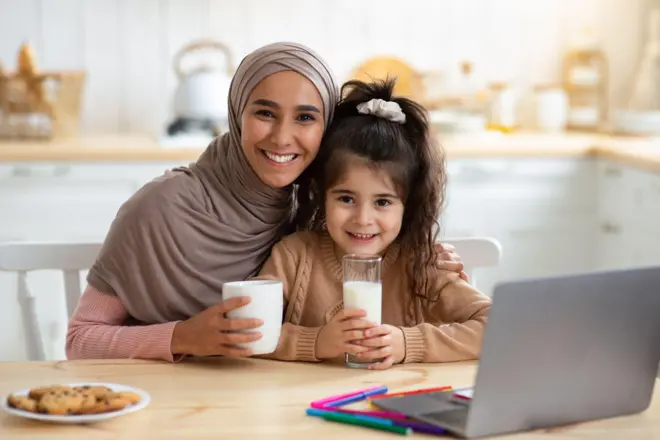 امرأة محجبة تجلس بجانب طفلة على طاولة في مطبخ، تمسك كل واحدة منهما كوب حليب، أمامهما حاسوب محمول، وقطع من البسكويت (الكوكيز) في صحن أبيض. 